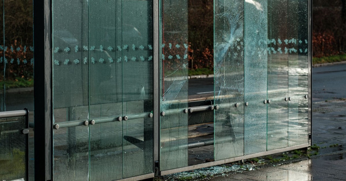 Damaged glass panels at a bus shelter on a rainy urban street, showcasing vandalism and city life.