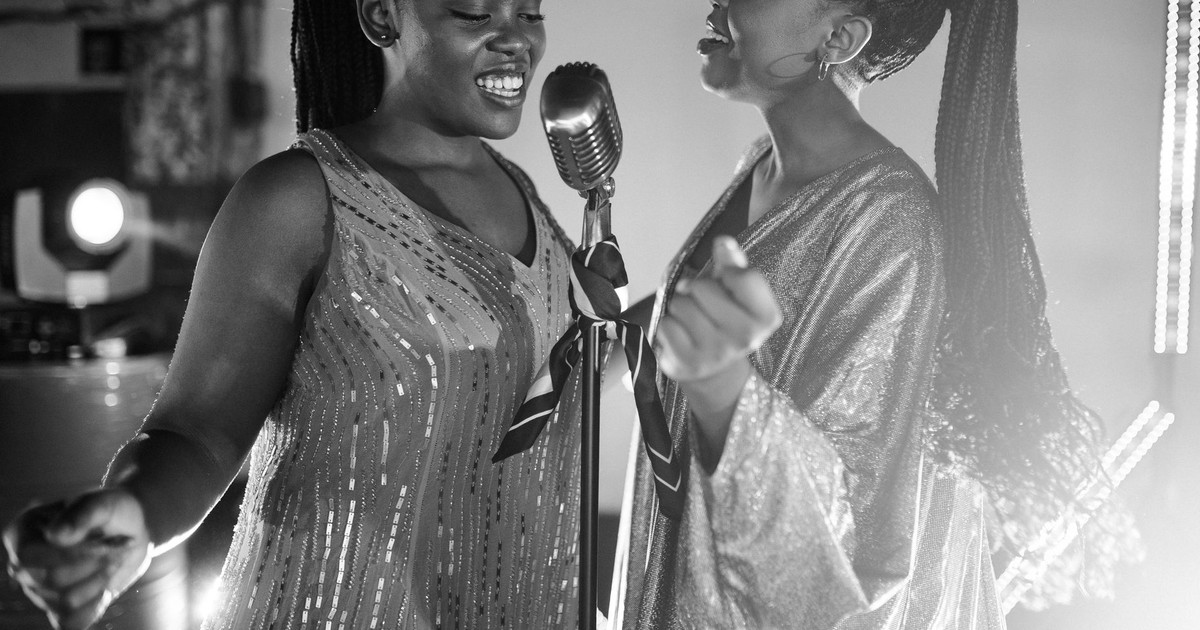 Two black women joyfully singing on stage with a retro microphone under bright lights.