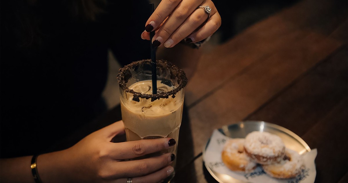 A woman stirring iced coffee in a dimly lit café setting, with powdered donuts on the side.