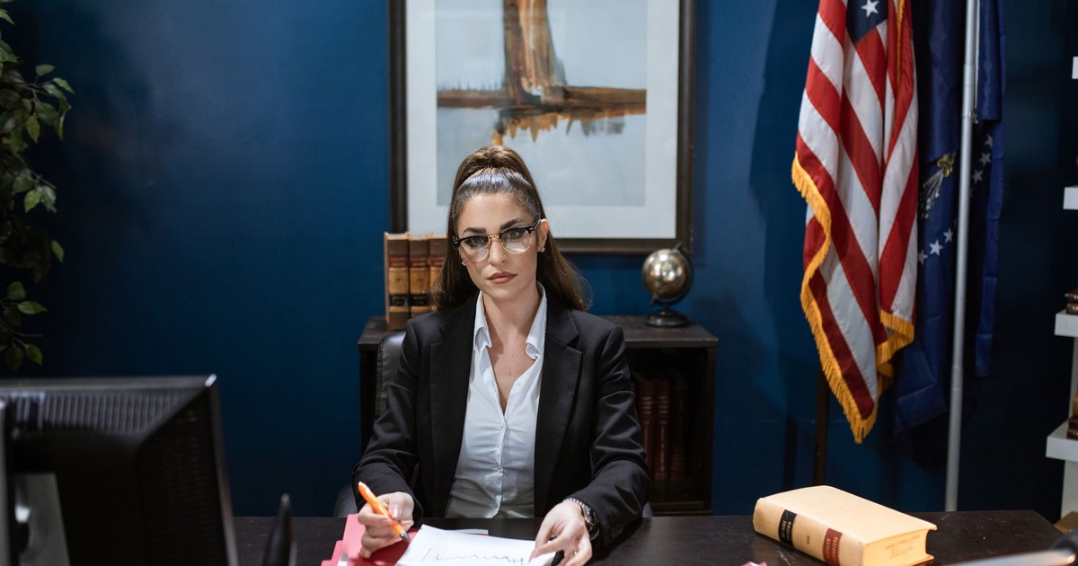 Focused woman in black blazer working on documents at desk with American flag in the background.