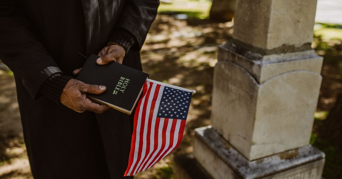 A person solemnly holds a Bible and American flag near a tombstone in a cemetery.