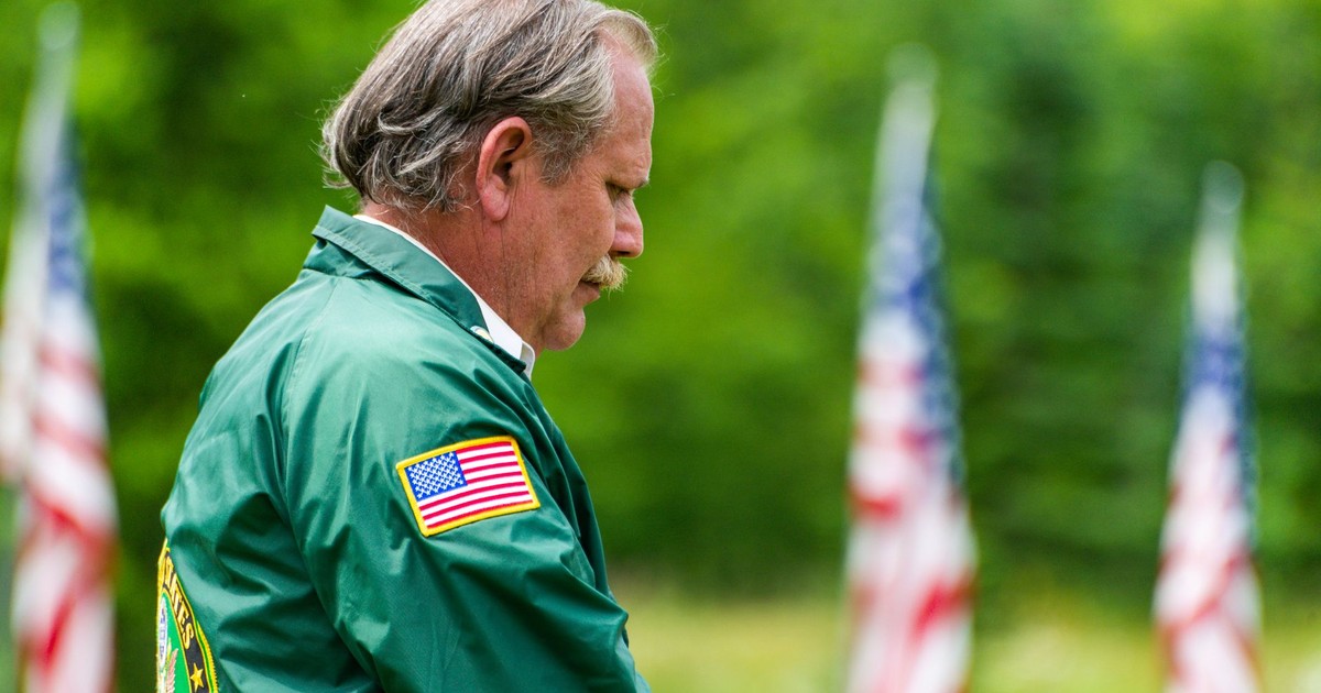 Senior adult standing outdoors, wearing a green jacket with USA flag, surrounded by American flags in a park setting.