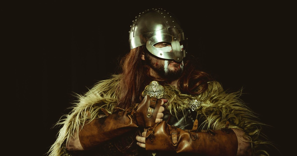 A viking warrior wearing a helmet and armor with fur covering against a dark background.
