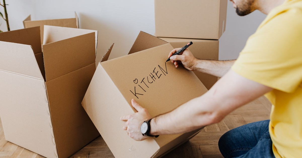 Focused young bearded male wearing yellow t shirt and jeans signing KITCHEN with marker on carton moving box while preparing for relocation in new home