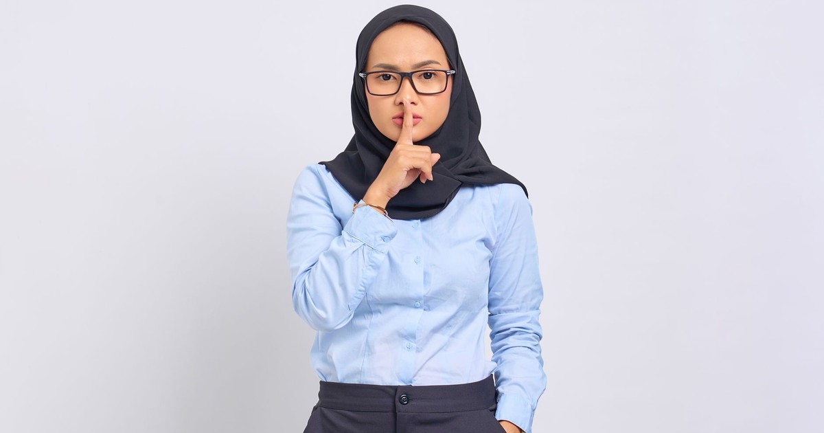 Asian Muslim woman in hijab making a quiet gesture with a serious expression, wearing eyeglasses against a white background.