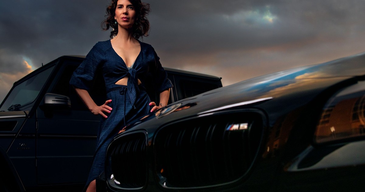Elegant woman stands confidently between luxury vehicles at sunset.