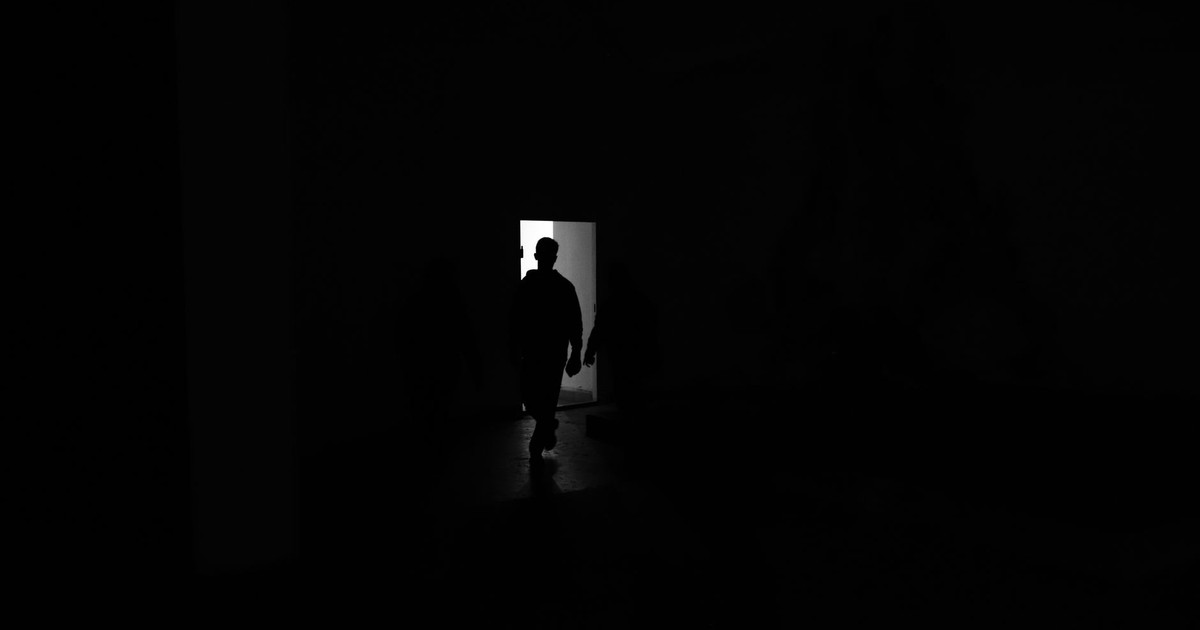 A shadowy figure stands silhouetted in a doorway, outlined by bright light.