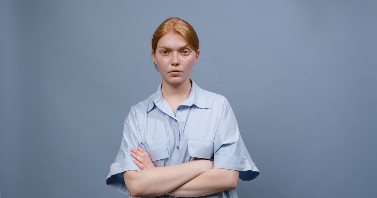 A young woman standing with crossed arms against a plain blue background, expressing annoyance.