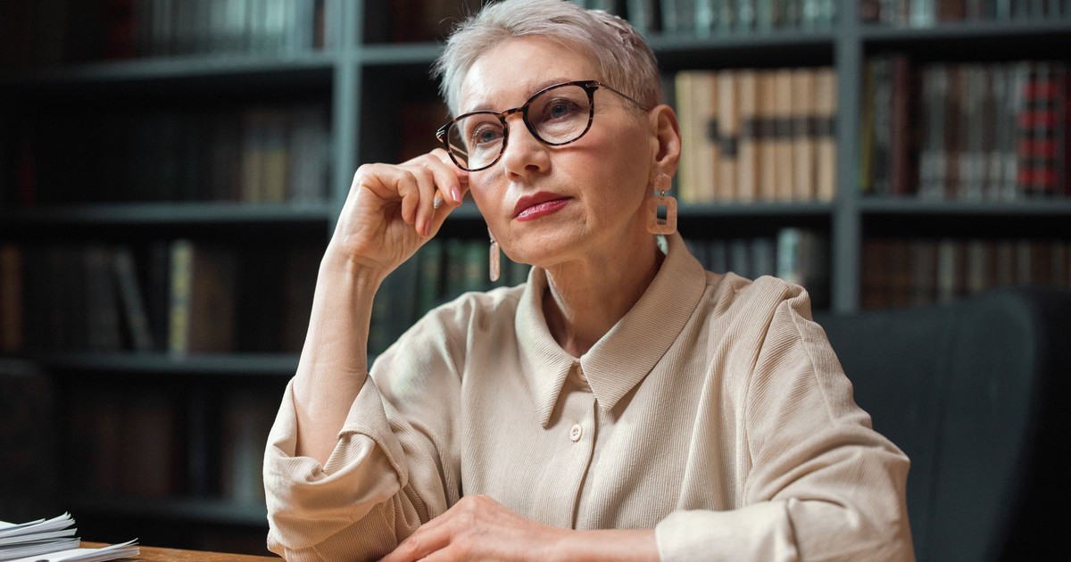 A reflective woman sitting at a desk in a library, surrounded by bookshelves, wearing glasses and earrings.