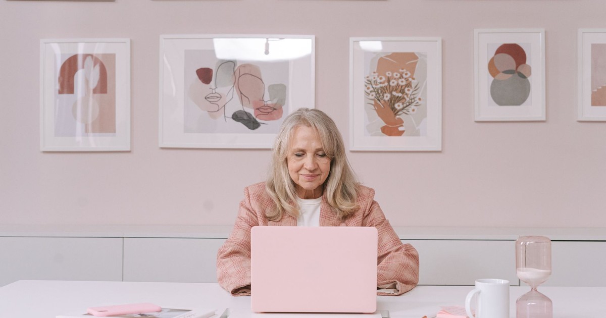 Elderly woman in pink coat working on a laptop at a white desk surrounded by artistic frames.