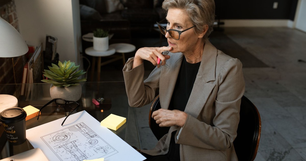 A senior architect in a gray blazer works thoughtfully at a home office with plans and a coffee.