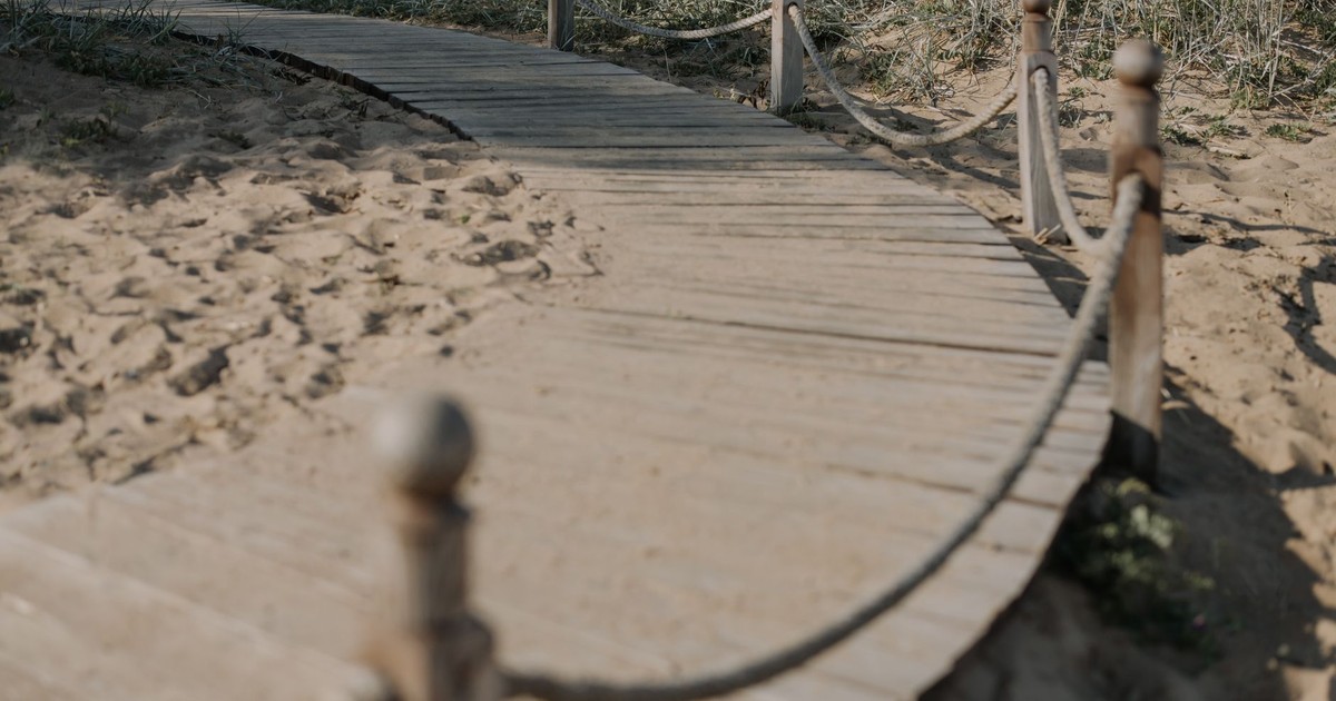 A close-up view of a curved wooden bridge on a sandy path surrounded by grass.