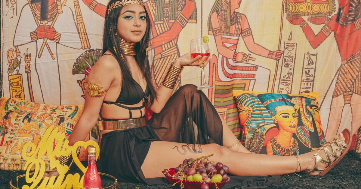 Stylized photo of a model in Egyptian-themed attire, holding a drink indoors.