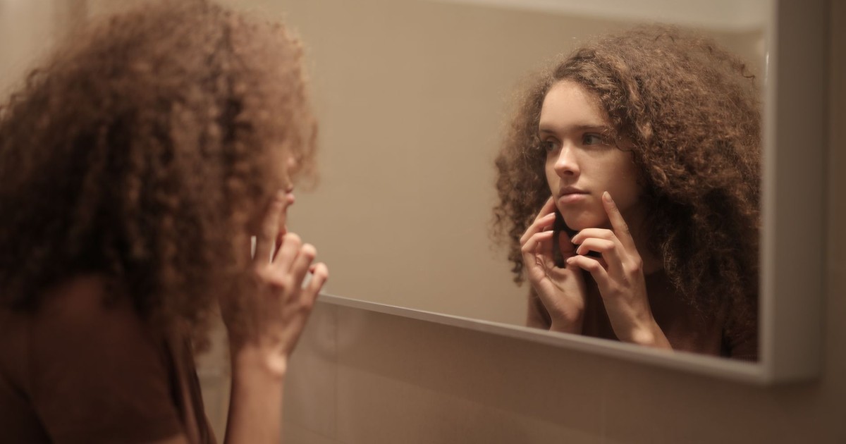 Side view of serious young lady with curly hair in casual clothes exploring face skin while looking at mirror in bathroom