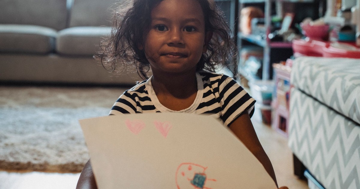 Cute ethnic girl in casual shirt demonstrating paper sheet with drawing while sitting in living room and looking at camera