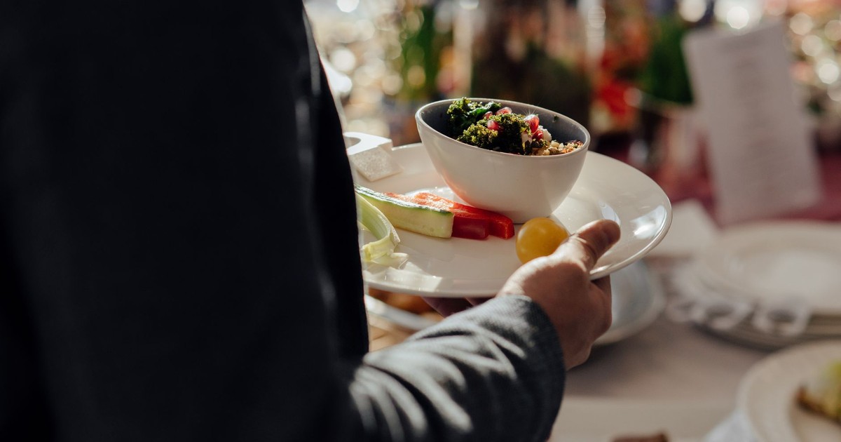 Crop faceless male with bowl of bulgur with spinach and pomegranate seeds on white plate with cut vegetables standing neat table on blurred background