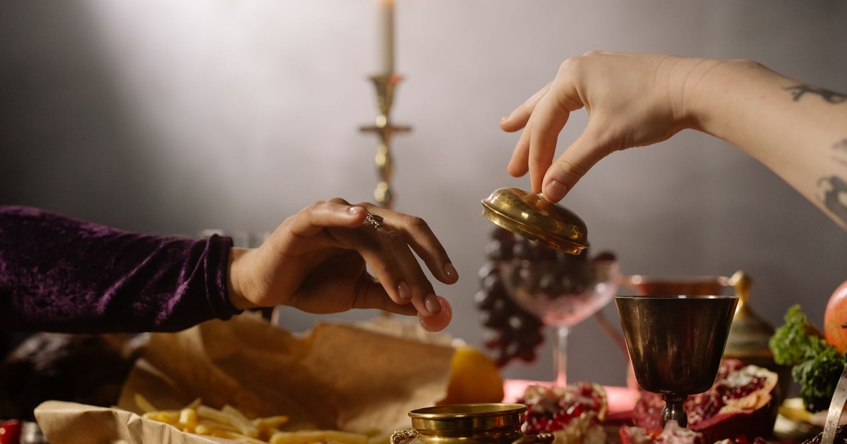 A moody candlelit dinner scene with hands reaching for golden containers amidst grapes and a pomegranate.