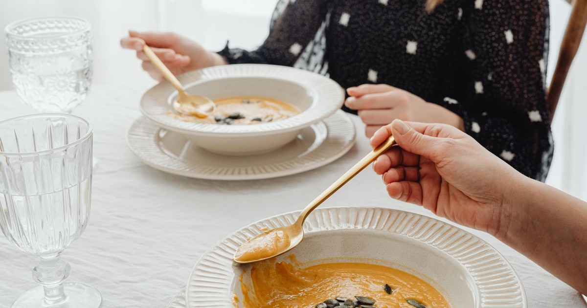 A close-up of hands enjoying creamy pumpkin soup at an elegant dining table setting.
