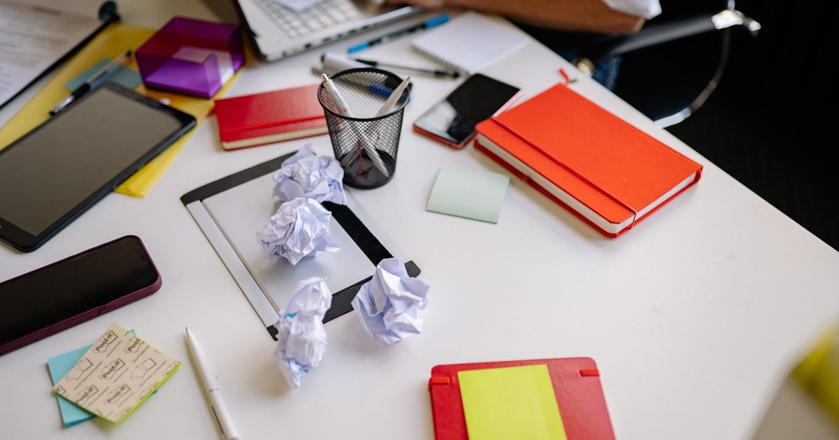 A cluttered office desk with crumpled papers, notebooks, and stationery.