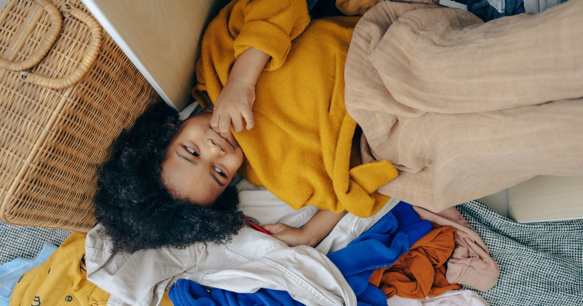 From above of playful little black girl smiling and lying on pile of various clothes after making mess in room