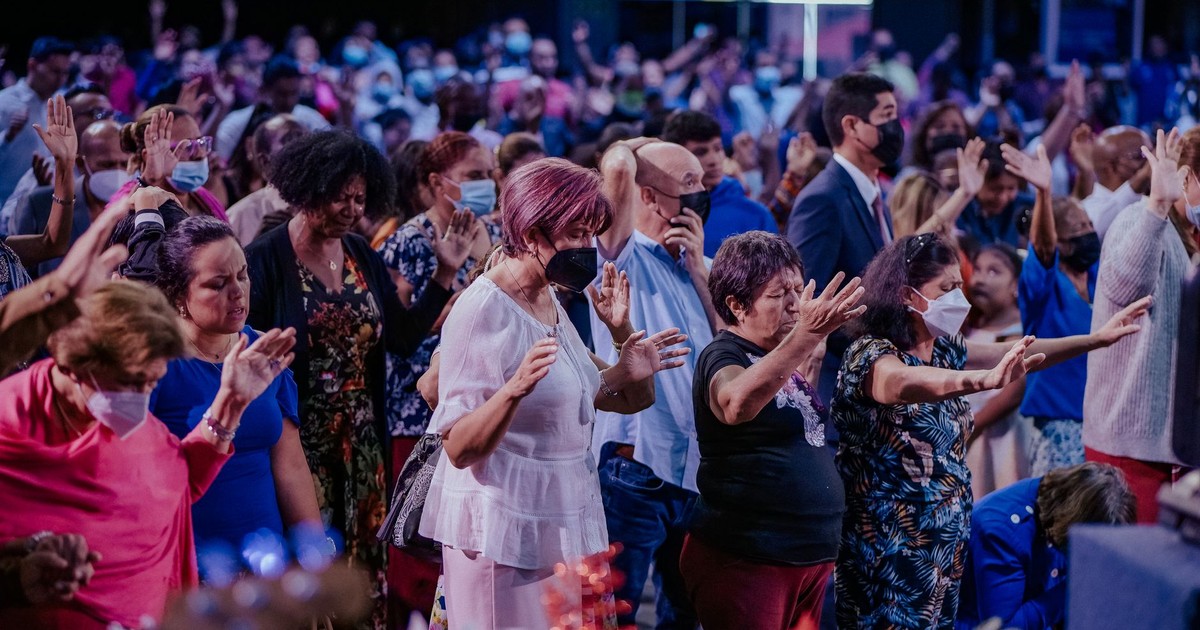 A large, diverse group of people praying and worshipping together indoors, wearing face masks.