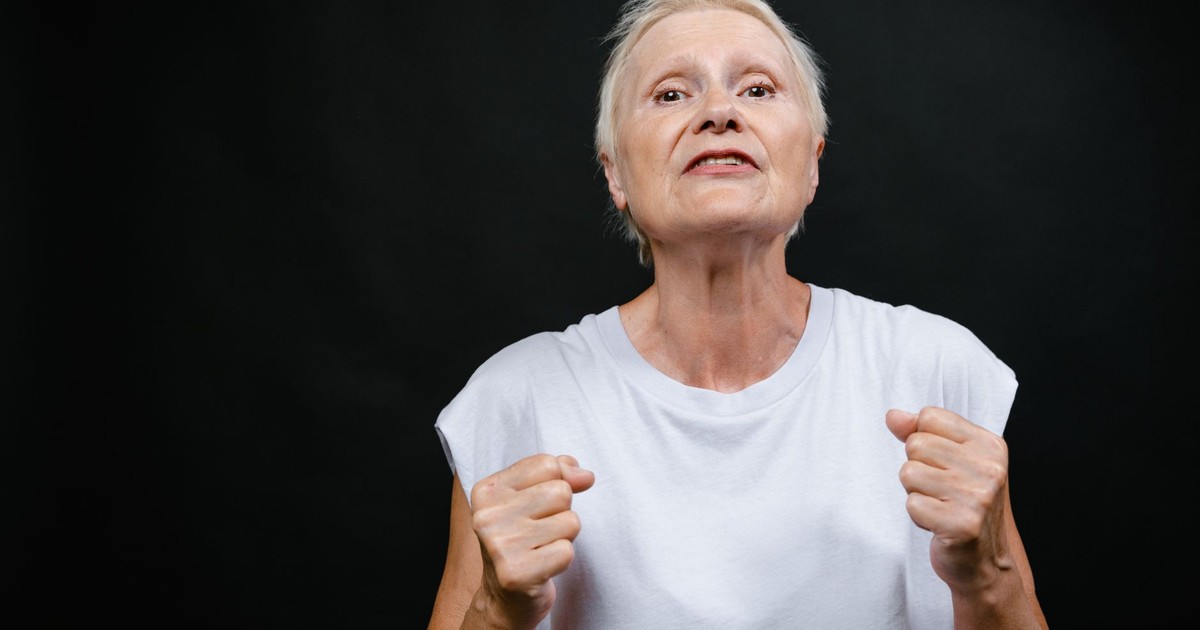 Determined elderly woman in white shirt clenching fists against black background.