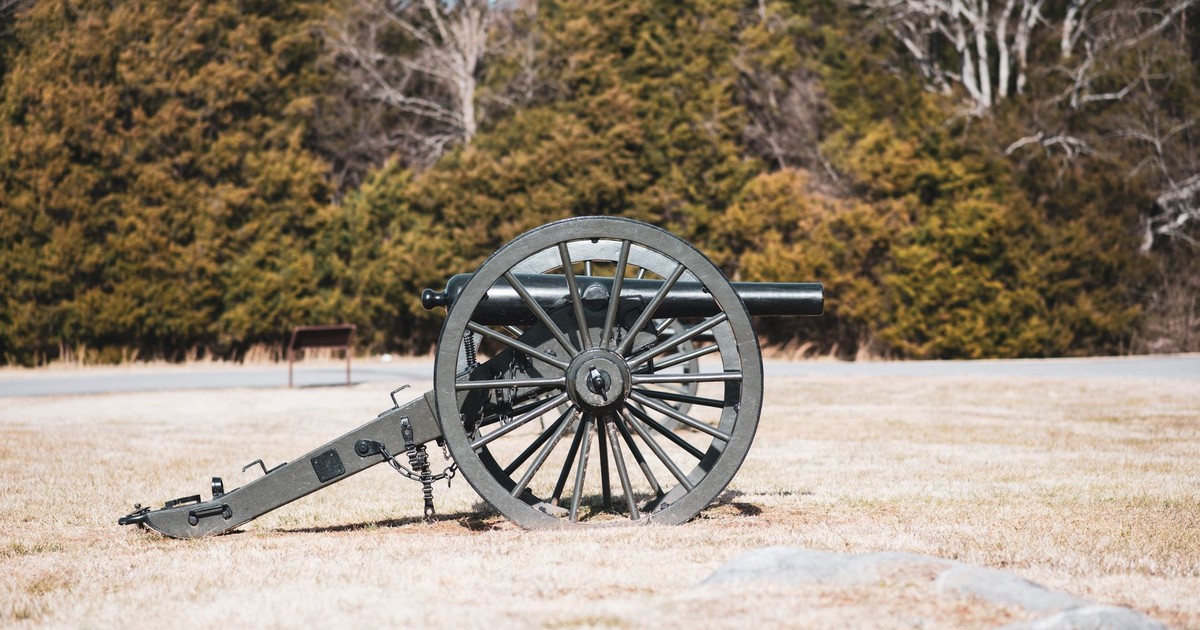 A historic cannon sits on grassy field under daylight, surrounded by trees.