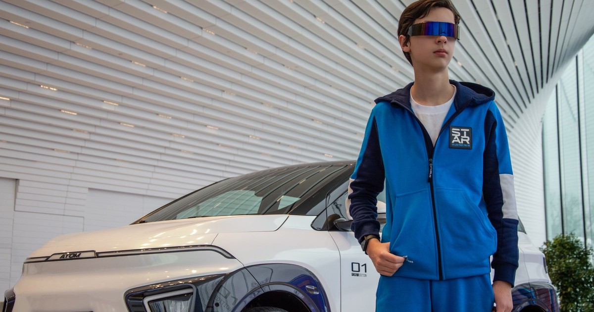 Teenager in blue tracksuit and futuristic glasses beside an electric car.