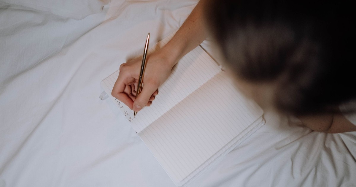 A person writing a to-do list in a notebook while sitting on a bed with white linen.
