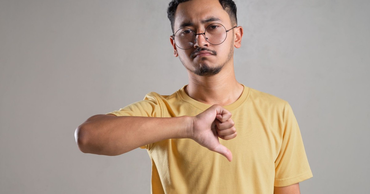 A man in glasses with a neutral expression giving a thumbs down gesture indoors.
