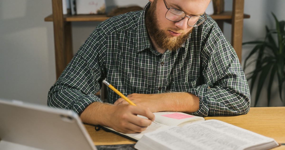 Man studying and taking notes with a Bible on a desk indoors.