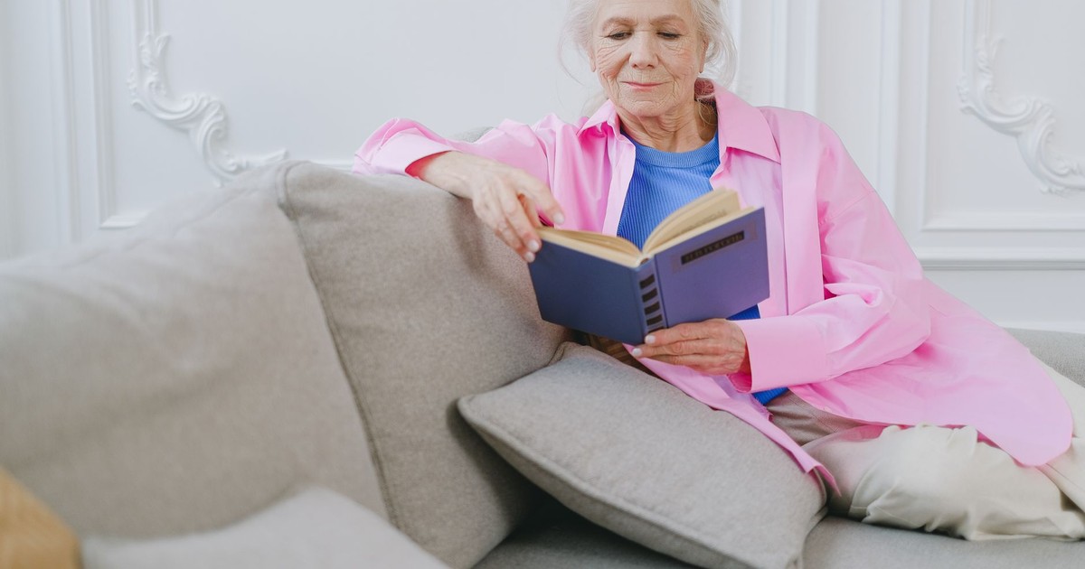 An elderly woman in a pink shirt enjoys relaxing on a couch while reading a book indoors.