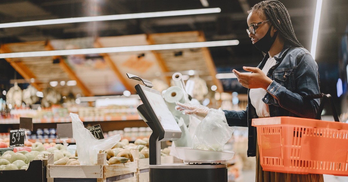 A woman with a mask shops and weighs groceries in a modern supermarket setting.