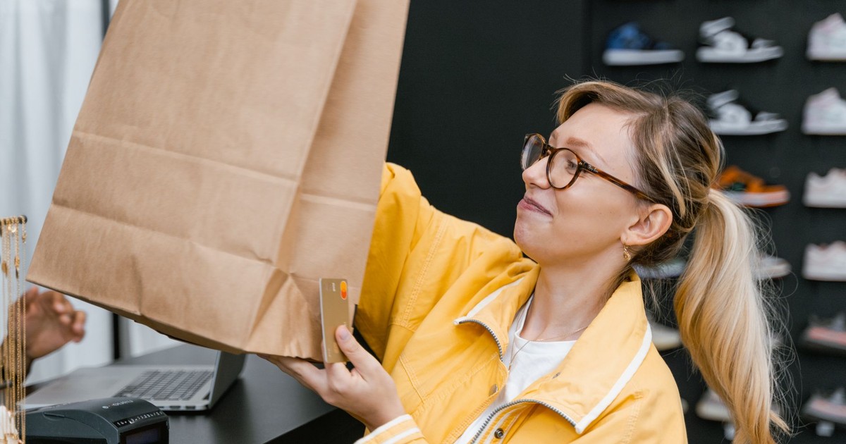 A woman in a store holding a brown paper bag and a credit card, smiling.