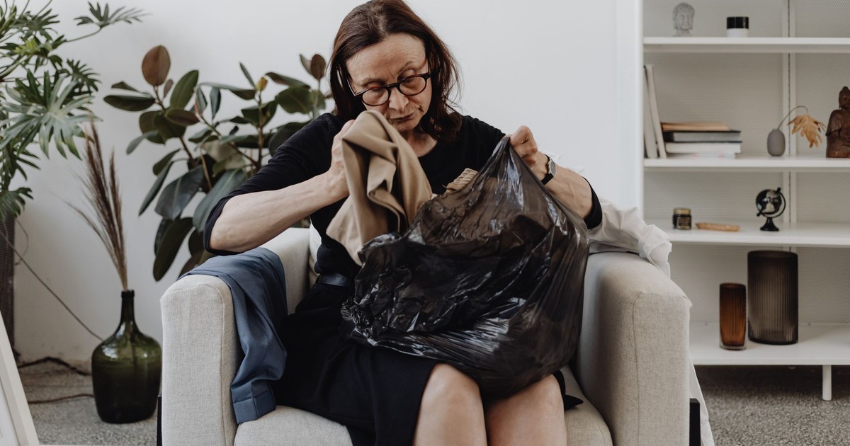 Senior woman seated indoors sorting clothes from a black bag, surrounded by plants and books.