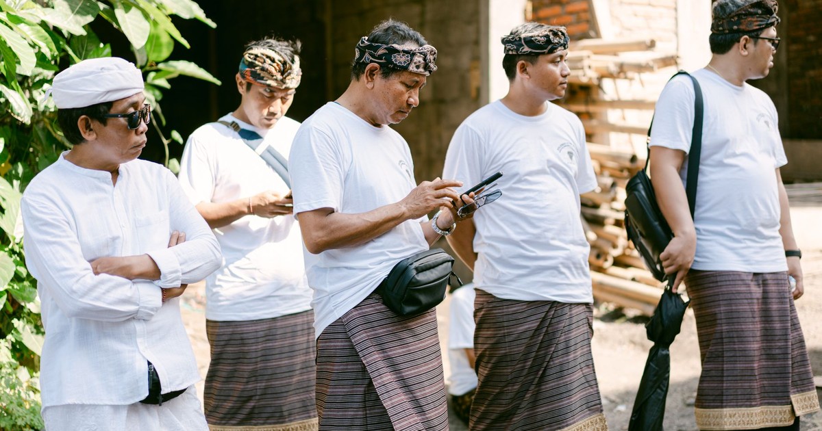 Group of men wearing traditional Balinese attire with smartphones, outdoors in daylight.