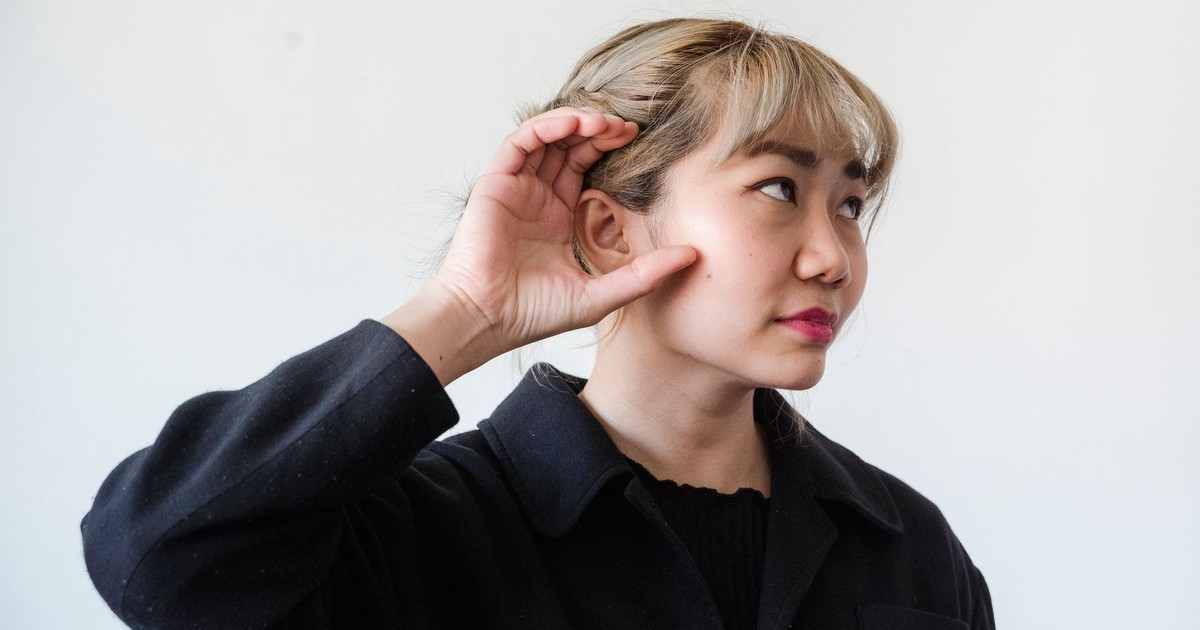 A thoughtful adult woman with blonde hair posing indoors, hand over ear.