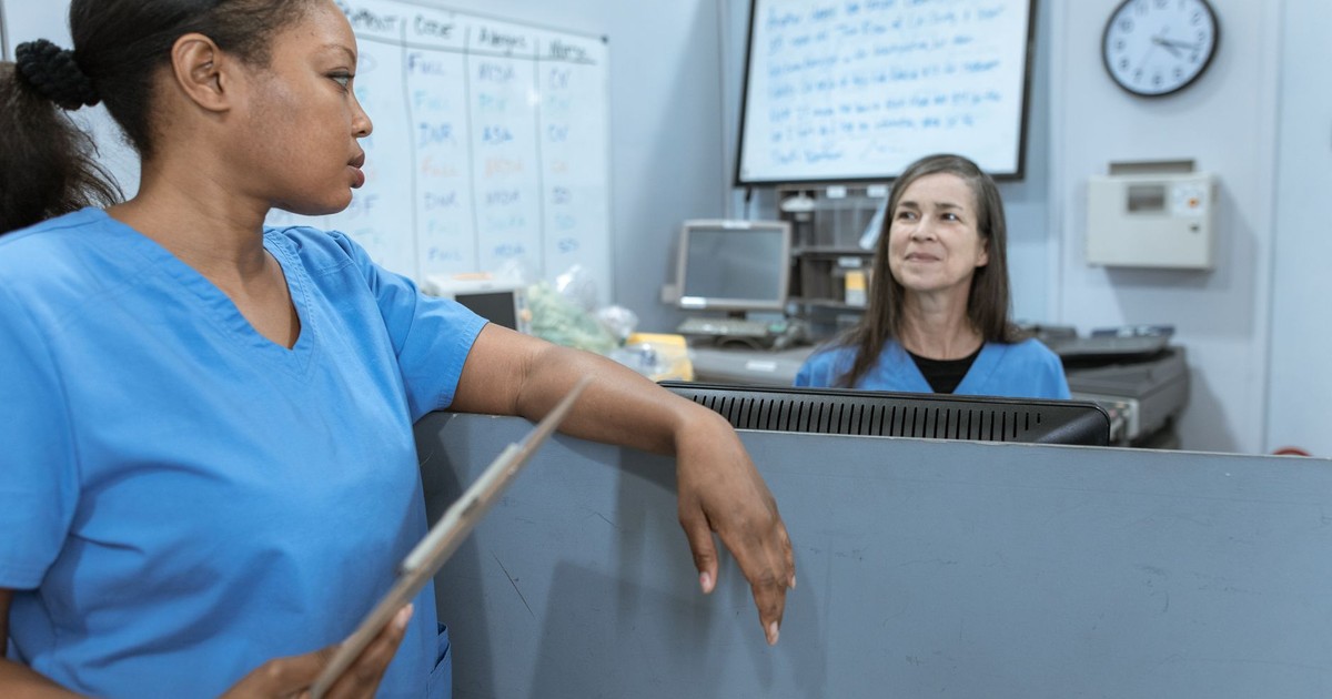 Nurses in scrub suits discussing medical tasks in a clinical environment.