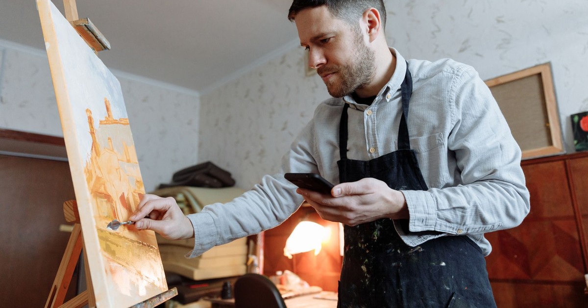 A male artist using a mobile phone while painting on a canvas with a palette knife in a studio.