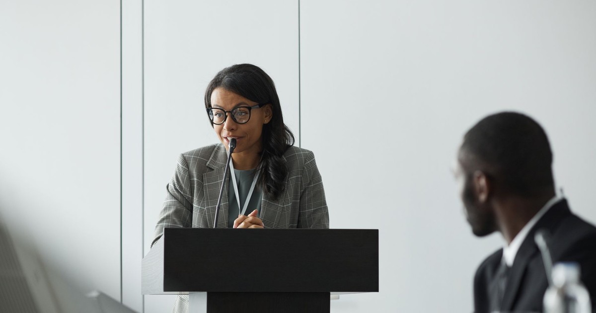 African American businesswoman delivering a speech at a conference podium with a colleague listening.