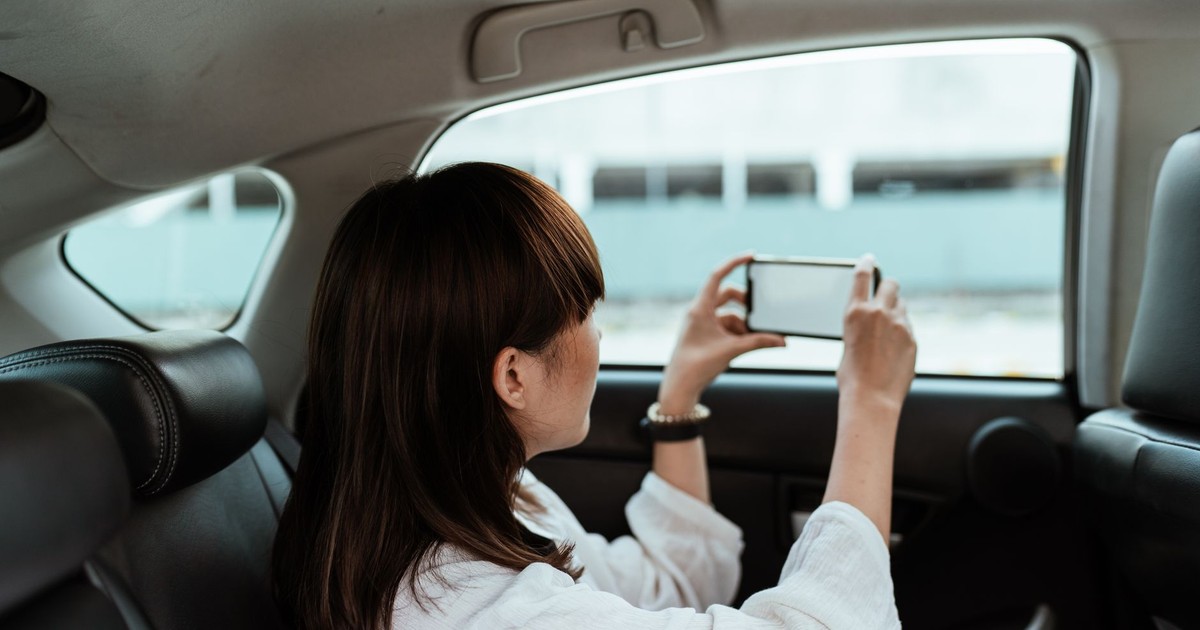 Side view of unrecognizable brunette in white shirt taking pictures on mobile phone with blank screen while riding in car backseat on daytime