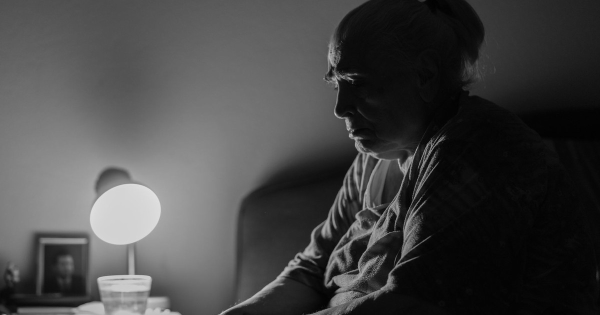 A contemplative senior woman sits by a lamp in a dimly lit room, evoking deep thought and solitude.