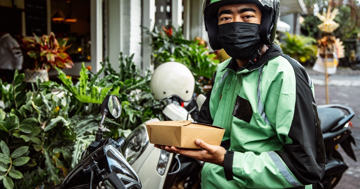 A delivery driver in green jacket holds food box while sitting on motorcycle.