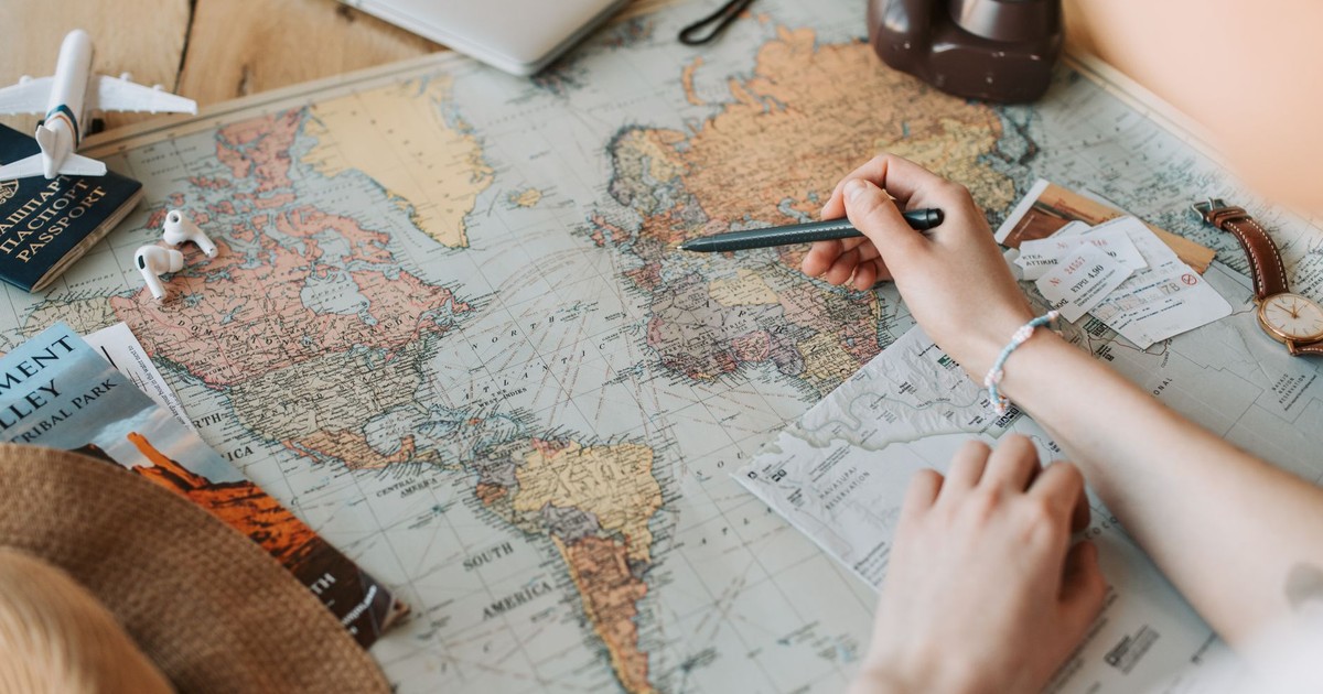 Hands marking destinations on a world map surrounded by travel essentials like passport and camera.
