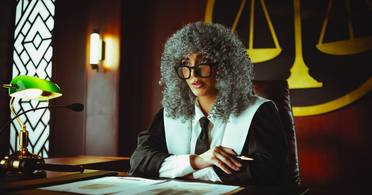Female judge in courtroom setting, sitting at desk with justice scales in background.