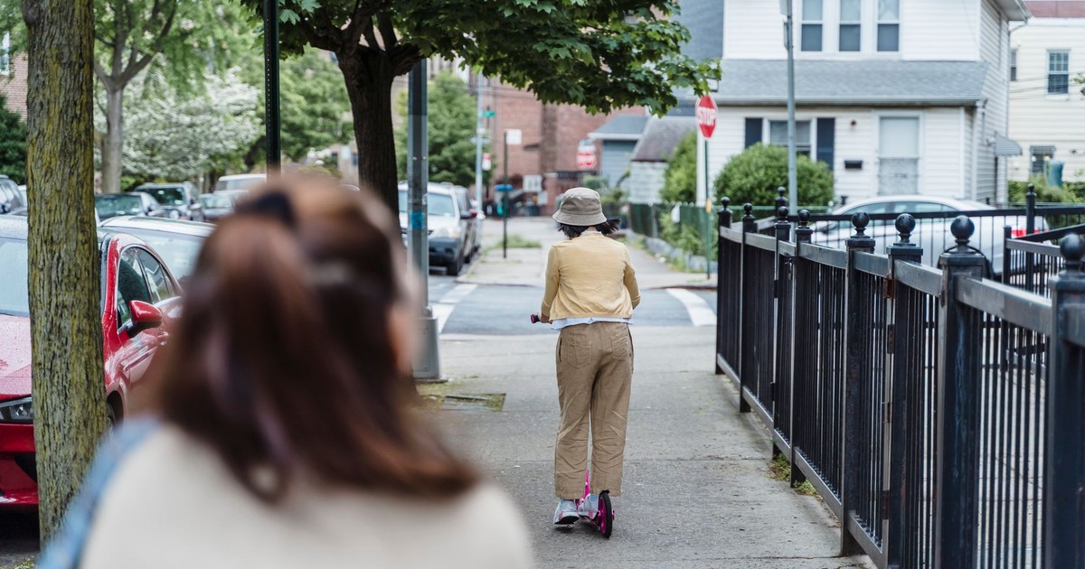 A young girl rides a scooter on a city sidewalk in a residential area.
