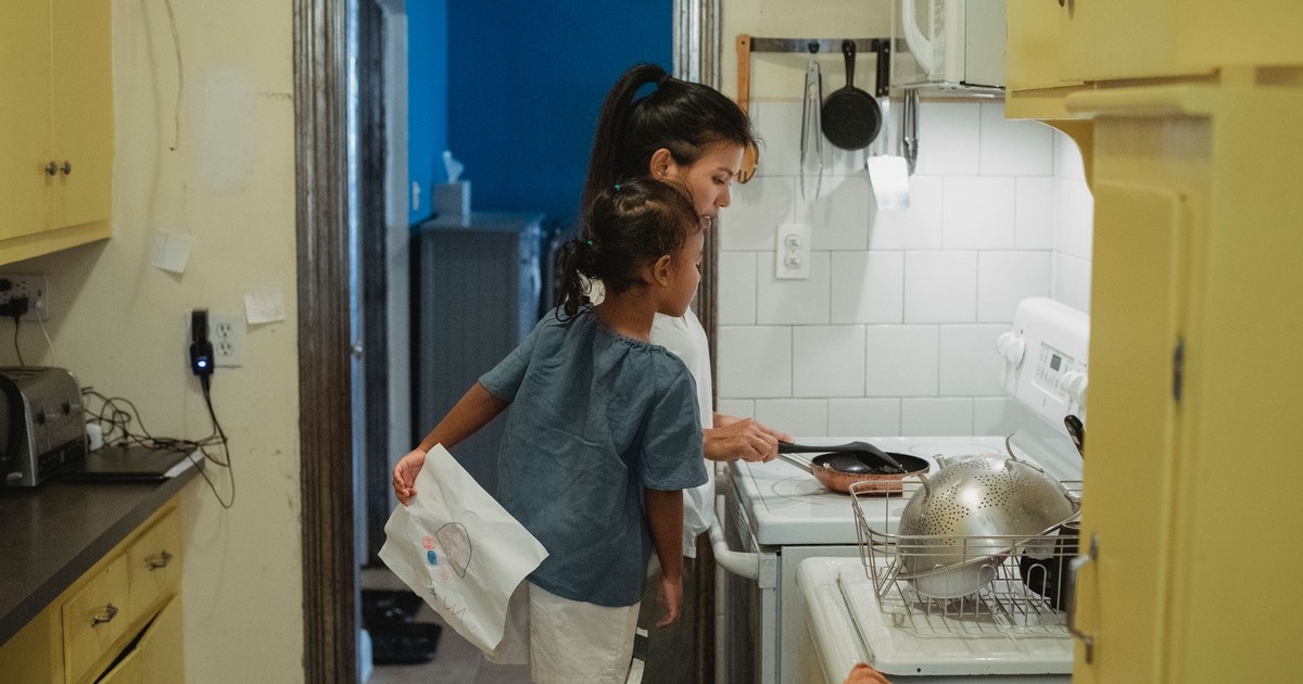 Side view of young Hispanic female and little girl in casual outfits standing near stove in kitchen and cooking together