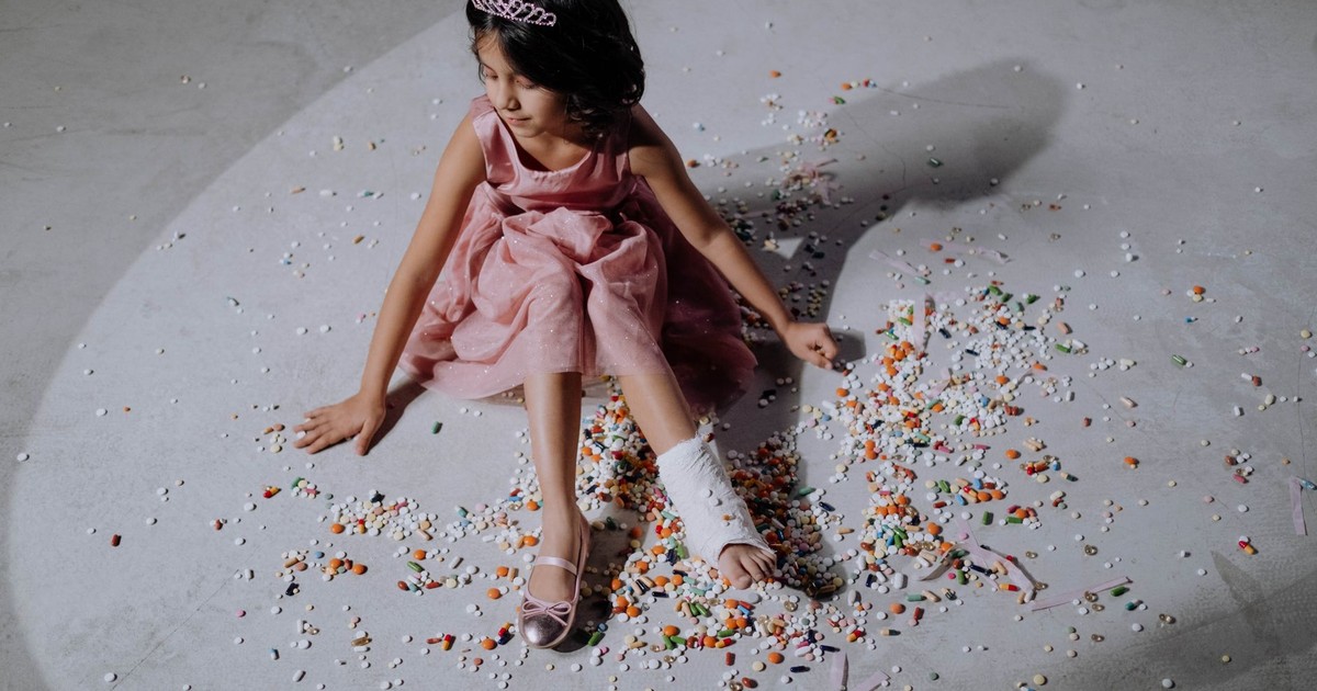 Asian girl in a pink dress with orthopedic cast, sitting on scattered pills.
