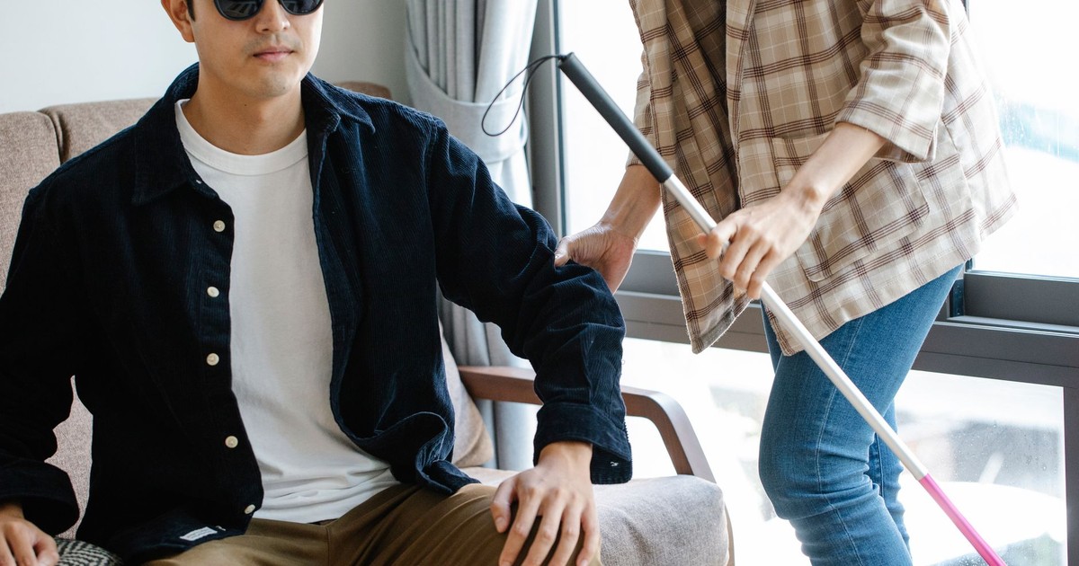 A visually impaired man receives support from a caregiver indoors, using a walking stick.