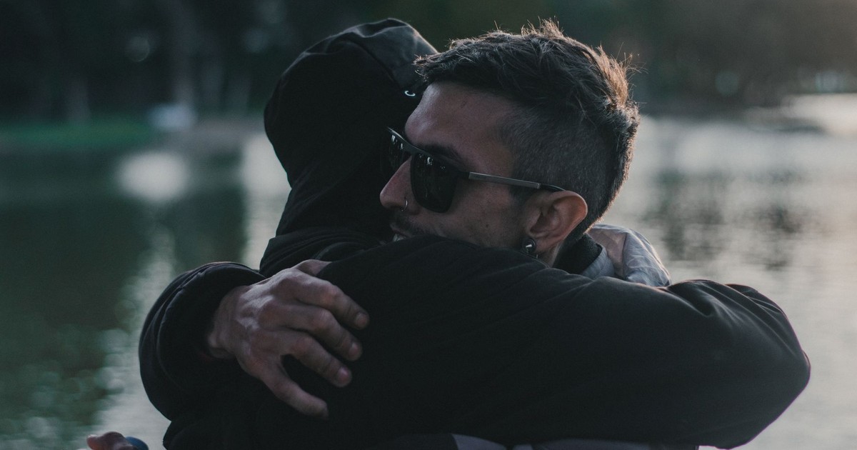 A heartfelt embrace between two adults near a calm lakeside during daylight.
